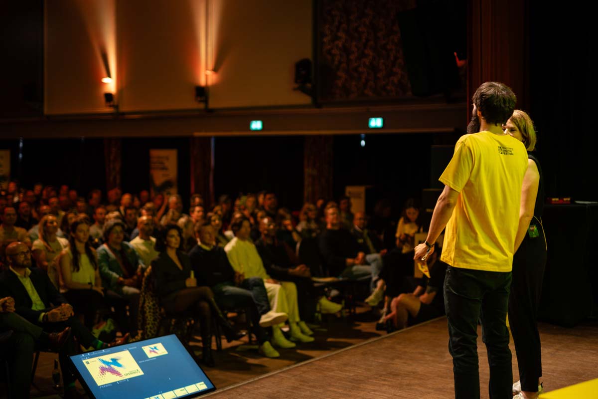 Speaker in yellow festival shirt in front of full auditorium.