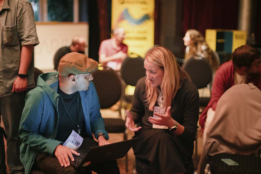 Two people in conversation, one with laptop, informal festival atmosphere.